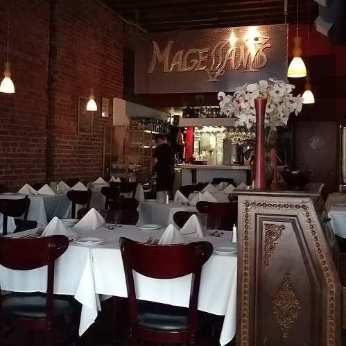 Interior of a restaurant with wooden tables, white tablecloths, and a bar in the background, featuring a floral arrangement.