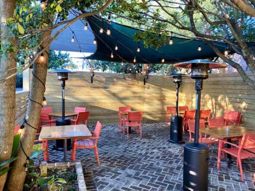 Outdoor dining area with red chairs, wooden tables, and patio heaters, shaded by a canopy and string lights.