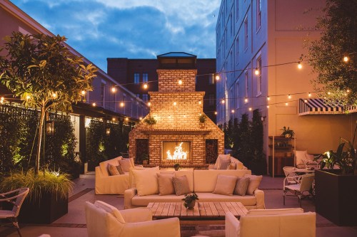 Cozy outdoor lounge area with a brick fireplace, string lights, and comfortable seating under a twilight sky.