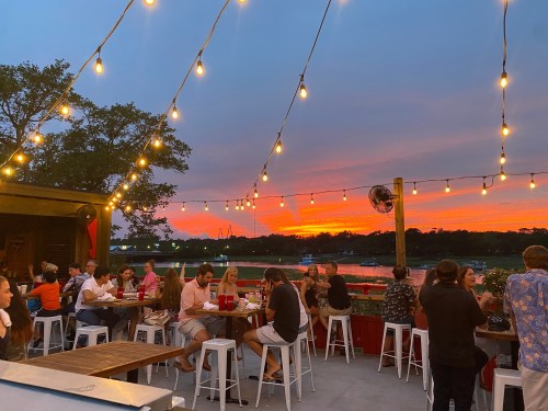 A lively outdoor dining scene at sunset, with string lights and people enjoying food and drinks by the water.