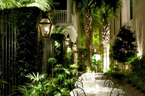 A beautifully lit outdoor dining area with tables, surrounded by lush greenery and elegant lanterns.