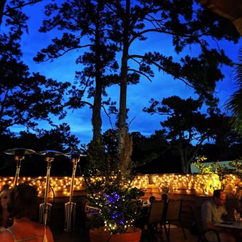 A cozy outdoor dining area at dusk, with string lights, patio heaters, and lush trees against a blue sky.