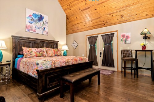 Cozy bedroom with a wooden bed, floral bedding, and a high ceiling, featuring a bench and double doors to the outside.