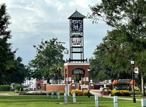 A clock tower stands in a park surrounded by trees and flowers, with buildings visible in the background.