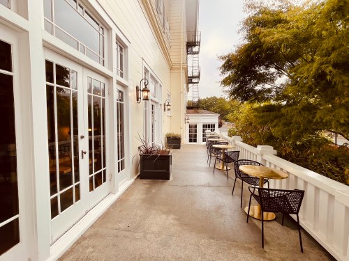 A walkway with white walls, large windows, and outdoor seating surrounded by greenery.