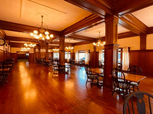 A spacious dining area with wooden tables, chairs, and elegant chandeliers, featuring warm lighting and polished floors.