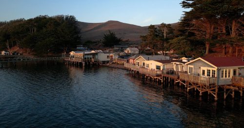 A serene waterfront scene with wooden houses on stilts, surrounded by trees and hills under a soft sunset glow.