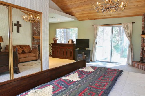 Cozy bedroom with a quilted bedspread, mirrored closet, and a view of a living area with a fireplace and large windows.