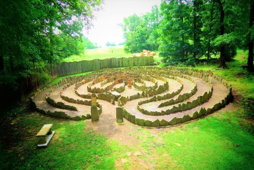 A stone labyrinth surrounded by trees, set in a grassy area with a wooden fence in the background.