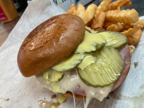 A close-up of a sandwich with ham, melted cheese, pickles, and a shiny bun, served with crinkle-cut fries.