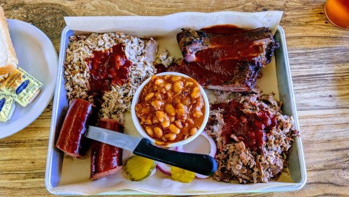 A metal tray with barbecue meats, rice, baked beans, pickles, and a knife, served on parchment paper.