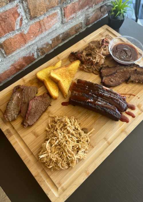 A wooden platter featuring various meats, including brisket, pulled pork, and cornbread, with barbecue sauce on the side.