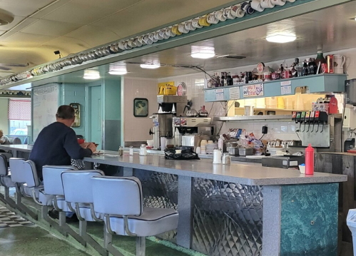 A diner interior with a counter, stools, and a man sitting alone, surrounded by condiments and a coffee machine.