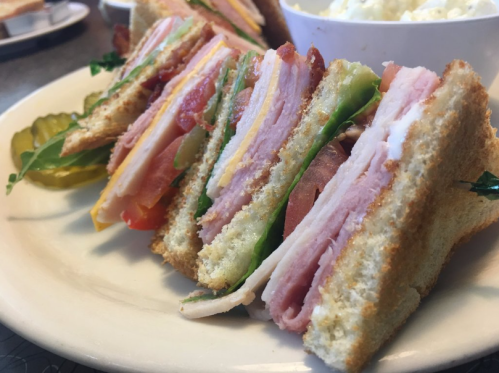 A close-up of a layered sandwich with ham, turkey, cheese, lettuce, and tomato, served with pickles on the side.