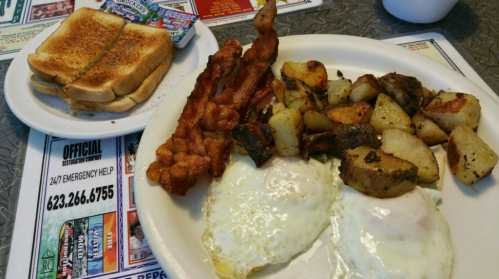 A plate with two fried eggs, crispy bacon, seasoned potatoes, and a side of toast on a table with menus.