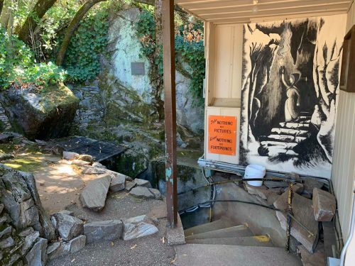 Entrance to a cave with a sign reading "No Flash Photography" and a mural of a figure in a cave. Surrounded by rocks and greenery.
