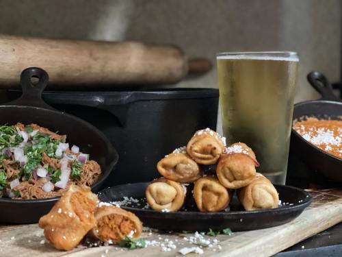 A platter of crispy tacos with a side of pulled meat, garnished with onions and herbs, alongside a glass of beer.
