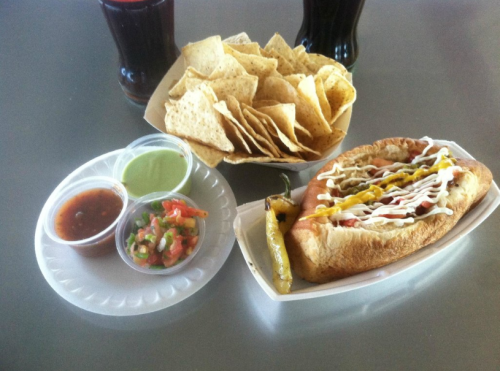 A hot dog with toppings, served with chips and two dipping sauces, alongside a small portion of salsa and guacamole.