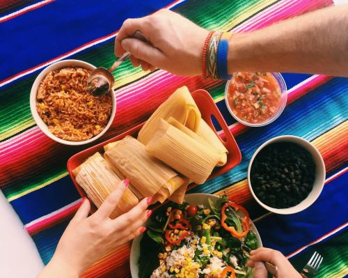 A colorful table setting with tamales, rice, beans, salsa, and a fresh salad, hands reaching for the food.