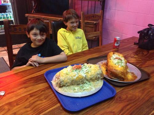 Two boys sit at a table with large plates of food, featuring oversized burritos topped with lettuce and tomatoes.