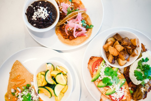 A vibrant spread of tacos, black beans, roasted potatoes, and sautéed vegetables on white plates.