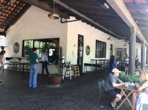 A sunny outdoor dining area with people seated at tables and a restaurant building in the background.
