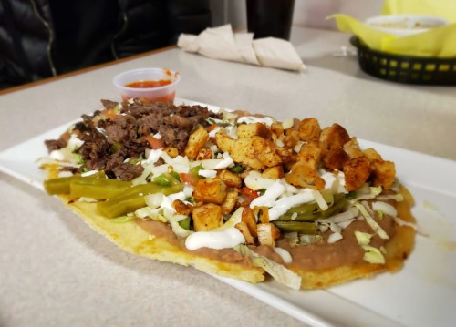 A large plate of nachos topped with beef, chicken, green peppers, lettuce, and drizzled with sauce, served with salsa.