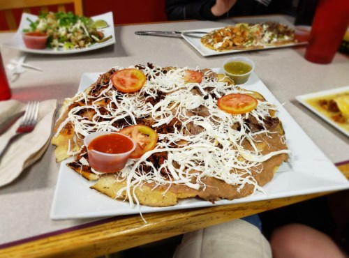 A large plate of nachos topped with shredded cheese, tomatoes, refried beans, and two sauces, served in a casual dining setting.