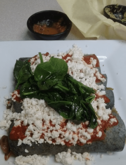 A plate of blue corn enchiladas topped with spinach, cheese, and red sauce, served with a small bowl of sauce.