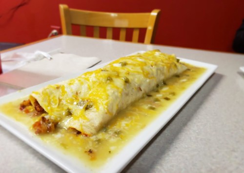 A large burrito topped with melted cheese and green sauce, served on a white plate on a table.