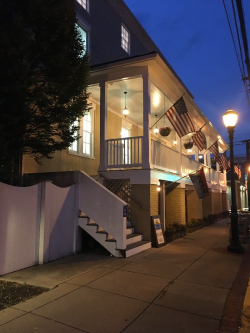 A charming building at dusk, featuring a porch with flags, illuminated windows, and a lamppost on a quiet street.