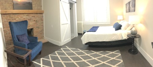 Cozy bedroom featuring a blue armchair, a bed with white linens, a brick fireplace, and a patterned rug.