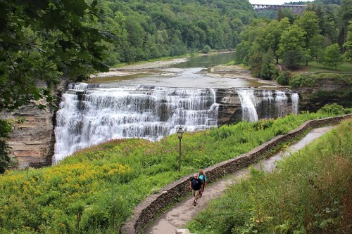 A hiker walks along a path beside a waterfall, surrounded by lush greenery and trees.