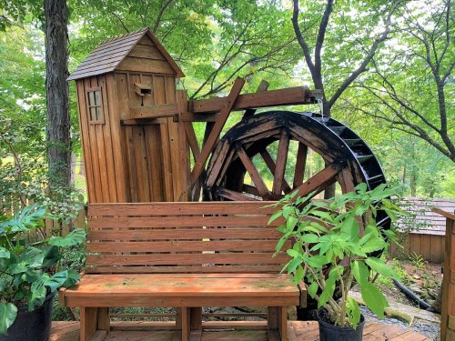 A wooden water wheel beside a small house structure, with a bench and potted plants in a lush green setting.