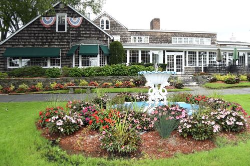 A charming building with green awnings, surrounded by colorful flower beds and a white fountain in the foreground.