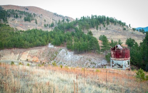 A weathered red water tank stands on a hillside surrounded by green trees and dry grass, with mountains in the background.