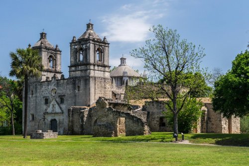 Historic stone mission with twin towers, surrounded by greenery and blue sky, showcasing weathered architecture.