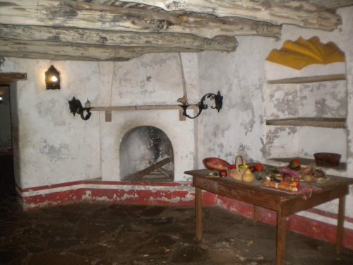 Interior of a rustic room with a stone fireplace, wooden shelves, and a table set with various fruits and food items.
