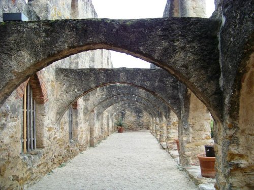Stone arches line a gravel path in a historic ruin, with potted plants and weathered walls creating a serene atmosphere.