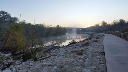 A serene river scene at dawn, with mist rising and a pathway lined by rocks and trees under a clear sky.