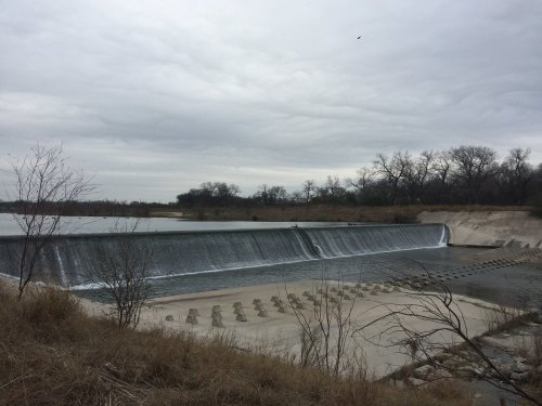 A serene view of a dam with cascading water, surrounded by bare trees and a cloudy sky.