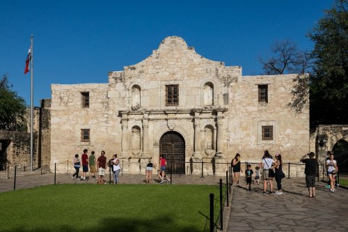 Visitors gather outside the historic Alamo building under a clear blue sky, with a Texas flag in the background.
