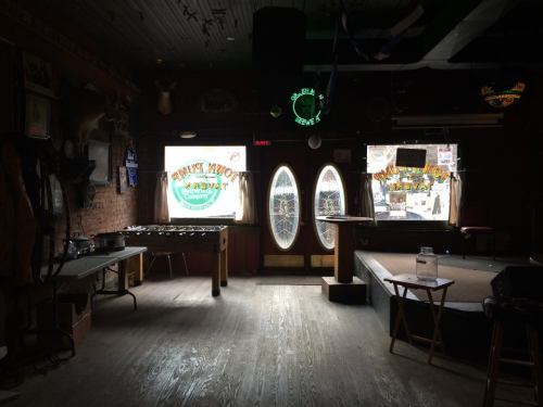 Interior of a dimly lit bar with wooden floors, a foosball table, and large oval windows.