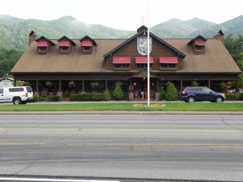 A rustic restaurant with a brown wooden exterior, red awnings, and mountains in the background. Cars are parked out front.