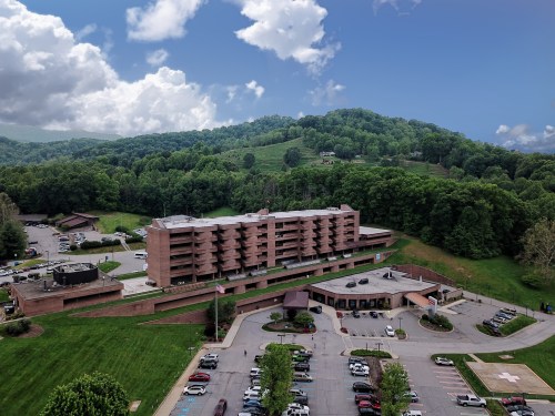 Aerial view of a hotel surrounded by green hills and trees, with a parking lot in the foreground under a partly cloudy sky.