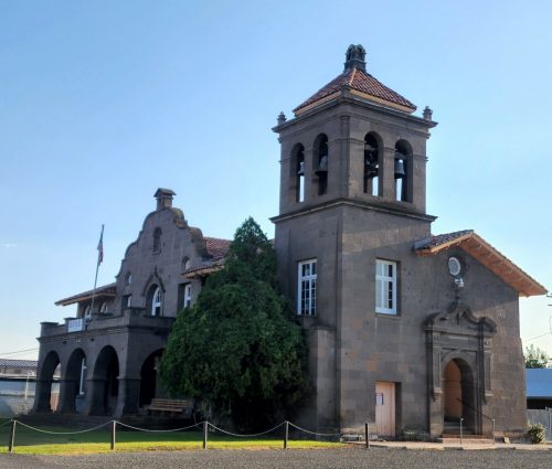 Historic stone building with a bell tower, arched windows, and a tiled roof, surrounded by greenery and a clear blue sky.