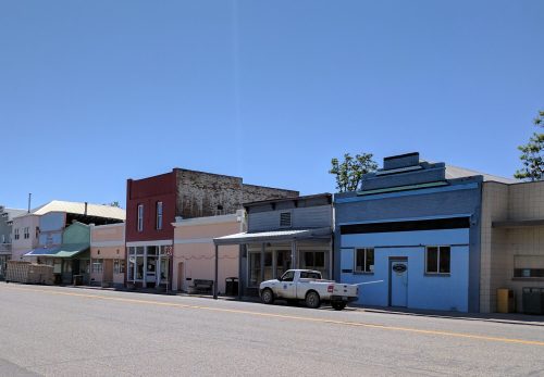 A quiet street scene featuring colorful storefronts and a parked white truck under a clear blue sky.