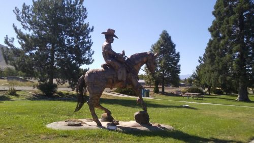 A wooden statue of a cowboy on horseback, surrounded by trees and grass in a park setting.