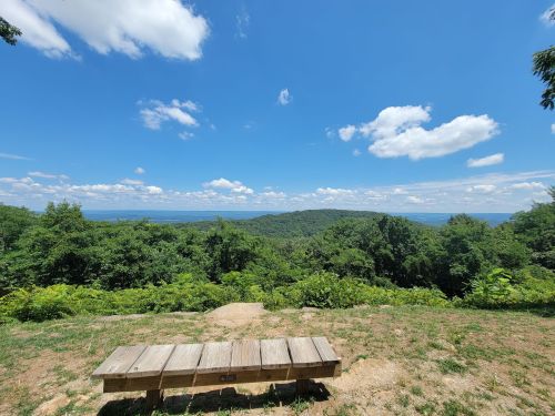 A wooden bench overlooks a lush green landscape under a bright blue sky with fluffy white clouds.