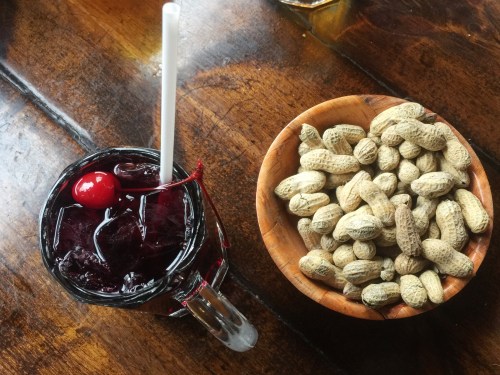 A glass of dark drink with a cherry and a bowl of unshelled peanuts on a wooden table.